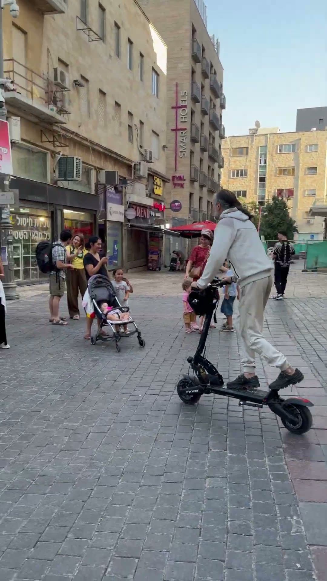 A man rides an electric scooter across the stone pedestrian mall on Ben Yehuda Street as families and children look on, central Jerusalem